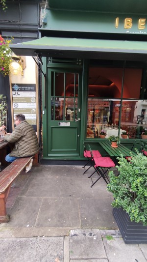 From sidewalk facing venue. Bench along left. Path ahead. Planters to right. Tables and chairs behind.