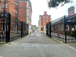 From street, facing venue. Path straight. Black metal barriers to left and right. Metal doors at end of path.