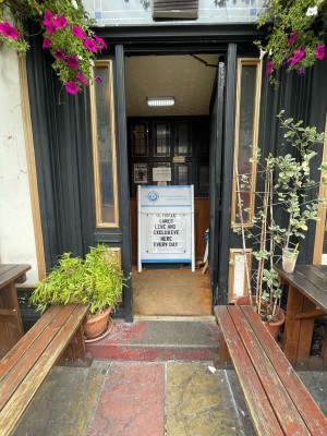 Path ahead leads to black and glass panelled door. Push from left of open. Planters to left and right of door. Outdoor seating on left and right.