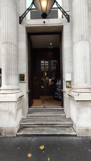 Facing venue entrance. Four steps up to brown wooden doors. White pillars on either side.