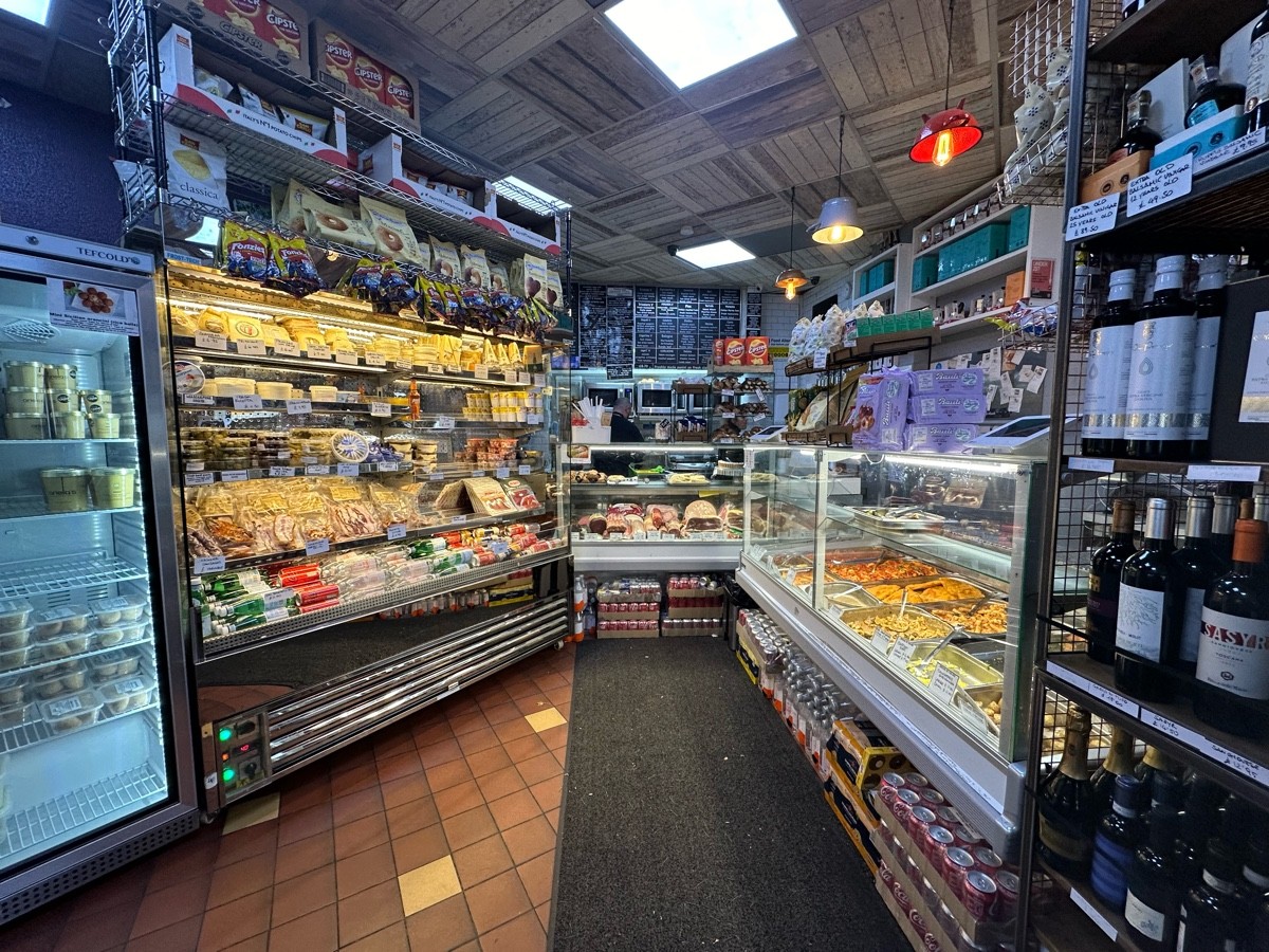 From high table, facing right. Fridge along left of path ahead. Retail shelves on right. Display counter beyond.