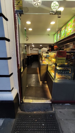 From sidewalk facing venue. Doormat on path followed by a short built-in ramp. Glass door on top of ramp. Push metal handle to right to open.
