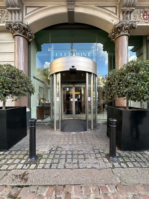 From pavement, facing straight. Opening ahead. Black pillars and planters to left and right of it. Path ahead leads to first doors.