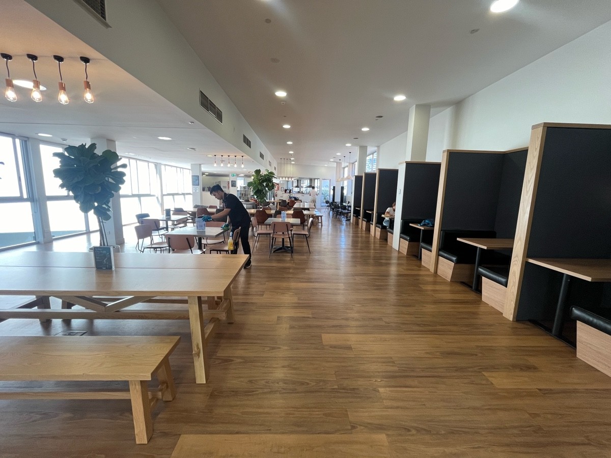 From path after pillar facing straight. Booths along right wall. Table with benches on left followed by tables and chairs. Path shifts right ahead.