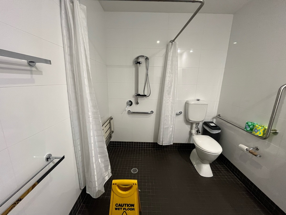 From door facing straight. Toilet on wall ahead. Shower on wall ahead in transfer space. Bench on left shower wall. Wet floor sign obstructing ahead.