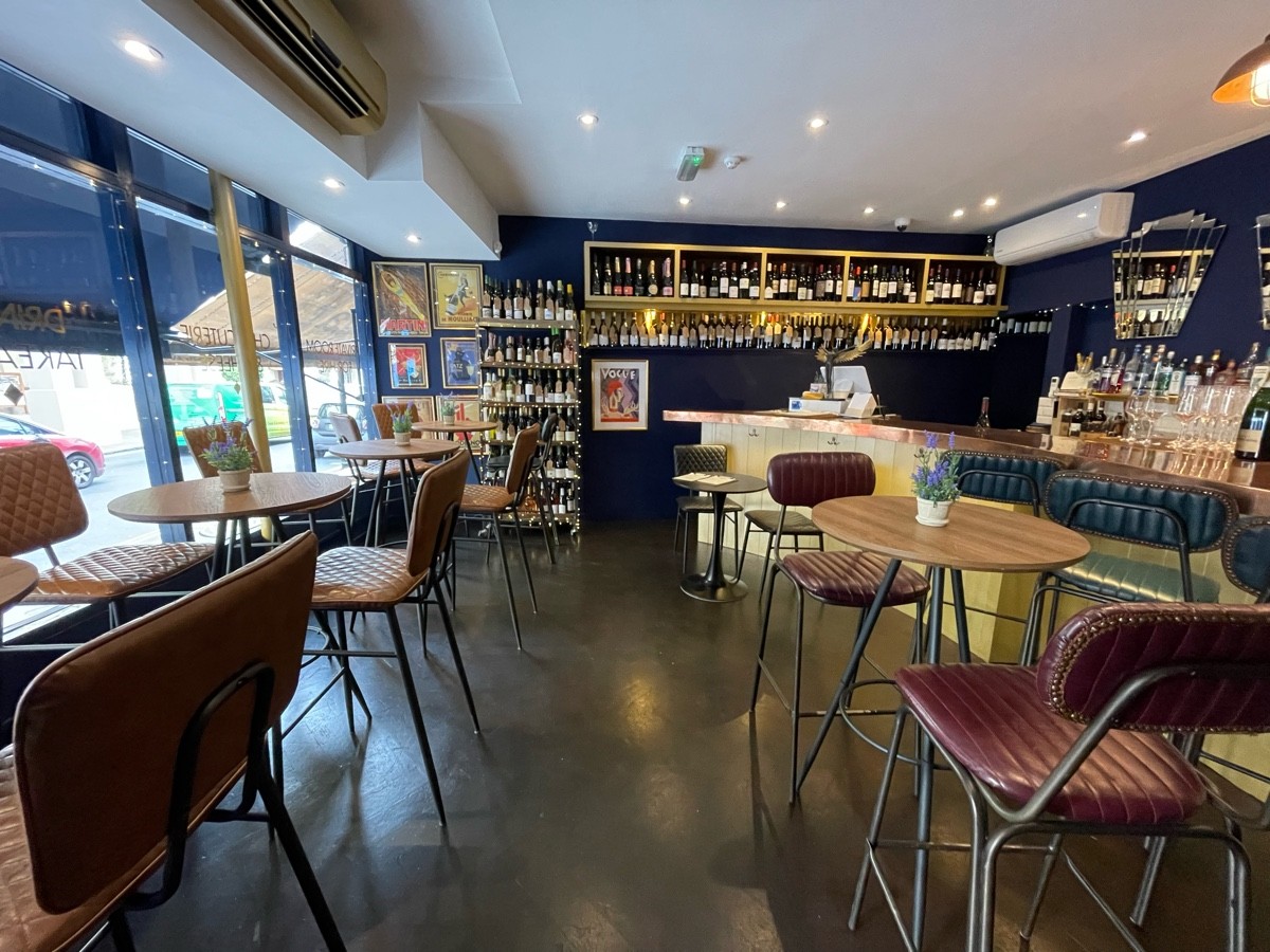 From entrance, facing left. High tables and stools along left and right. Path ahead. Bar along back right.