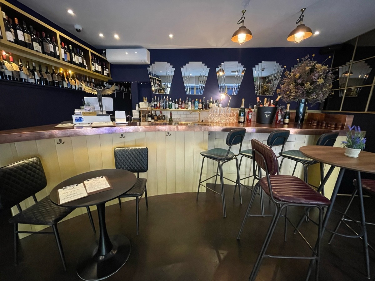 From end of tables and stools, facing right. High tables and stools ahead. Bar with stools along back wall.