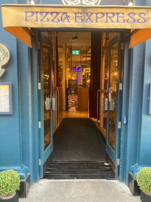 From side road, facing venue. Double glass door with blue frame ahead. Push open via metal handles in center. Blue doorbell to the right of doors.