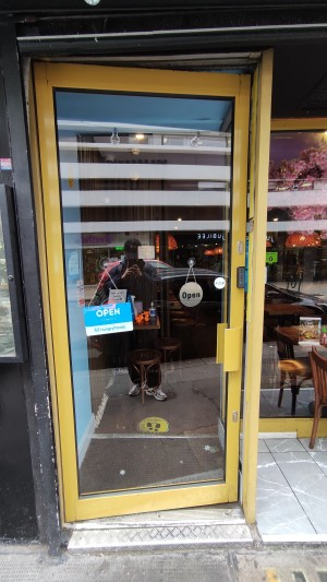 From sidewalk facing venue. One step up to glass door with yellow frames. Push yellow handle to right to open. Metal bump at foot of door.