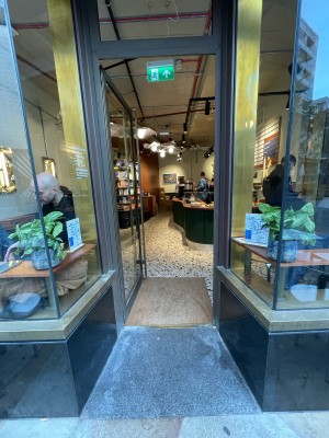 Glass door in metal frame. Push vertical golden handle on right to open. Glass walls to either side before entrance.