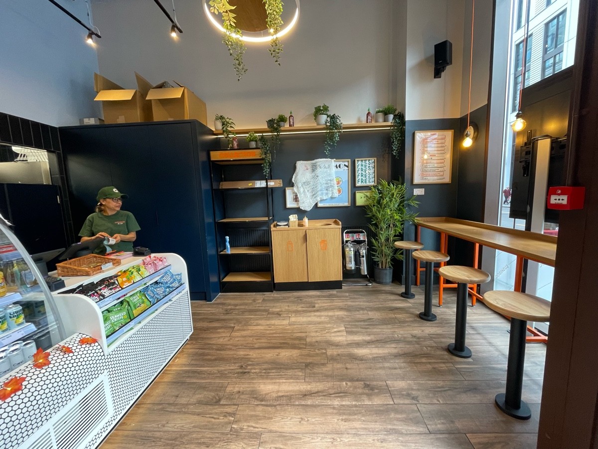 From entrance facing right. Counter along left. Pick up shelf and bin at back. High table with fixed stools along right.