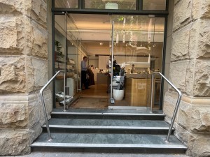 Glass double doors. Push silver vertical handles in centre to open. 3 steps up to doors. Metal handrail either side. Doormat after doors.