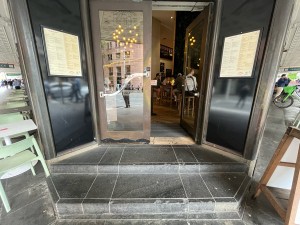 Glass double doors with brown metal frame. Push from middle to open. Two steps up to door. Doormat after door.