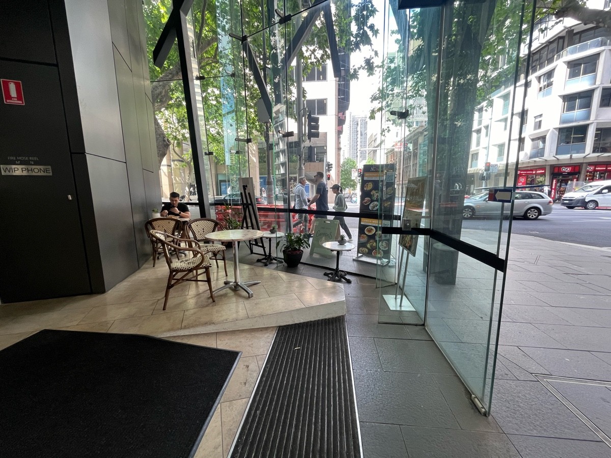 From before ramp facing right. Tables and chairs ahead to left and on right wall. Glass divider obstructing to right. One step up to seating on left.