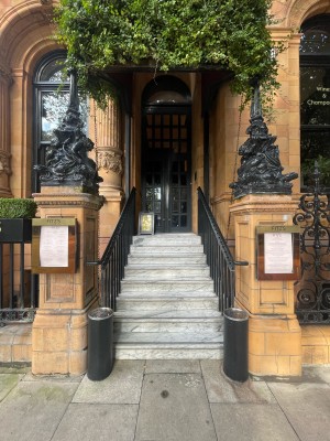 From street, facing venue.  Staircase with black metal handrails on both sides ahead. Ashtray stands to left and right. Wooden pillars beyond it.