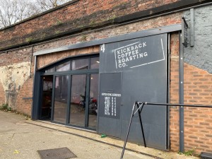 The image shows the outside of Kickback coffee. There is a slope down to the doorway. The front of the building is mostly glass.