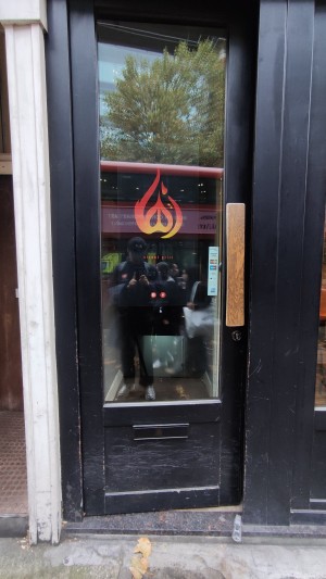 From sidewalk facing venue. One step up to glass door with black frames. Push wooden pad to right to open.