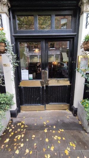 From sidewalk facing venue. Path ahead to glass doors with black frames. Push gold pads in centre to open. Planters on either side of doors.