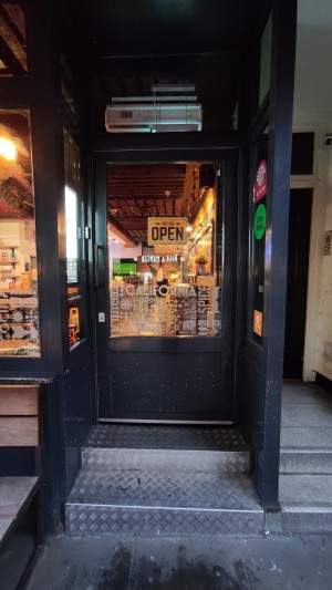 From sidewalk facing venue. Two steps up to glass door with black frames. Push black handle to left to open.