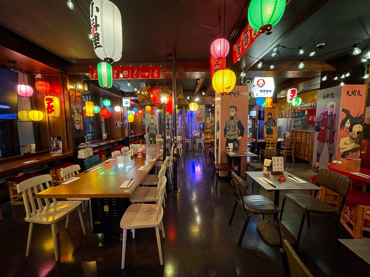 From end of long table with pillars, facing straight. Long table and chairs along left. Tables and chairs along right.