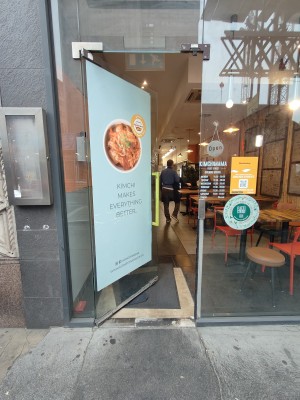 Facing glass door. Push yellow metal handle on right to open.