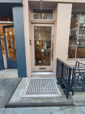 Path ahead to entrance. Glass door in pink wood frame. Push golden metal handle on right to open. Doorbell on left of door. Black handrail on right.