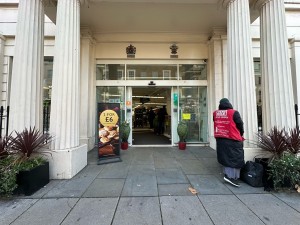 From pavement, facing venue. Opening ahead. White pillars and planters on left and right. Standing sign to back let before doors.