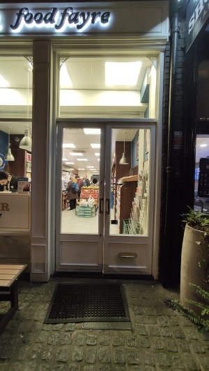 From pavement facing venue. Path ahead with doormat at the end. Glass doors with white frames at the back. Push gold handles in centre to open.