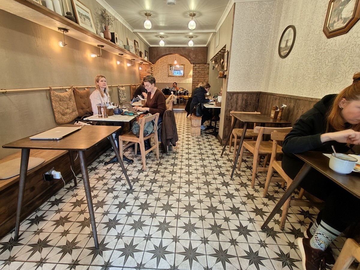 From end of display counter facing straight. Half booth along left. Path ahead to back area and toilet. Tables and chairs on right. Half booth behind.