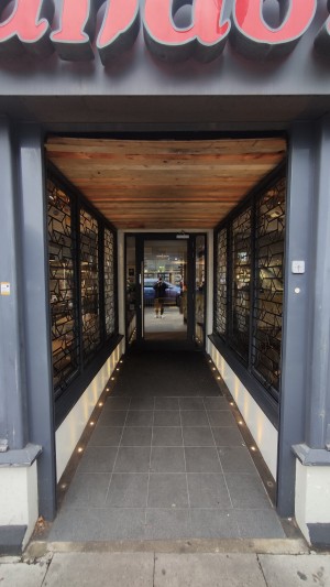 From pavement facing venue. Open doorway ahead. Built in ramp in centre. Glass walls on left and right.