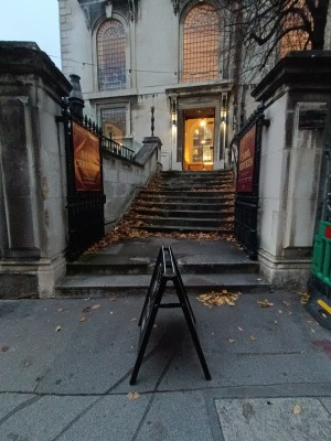 From street, facing venue. A-frame board ahead. Open black, metal gates ahead.