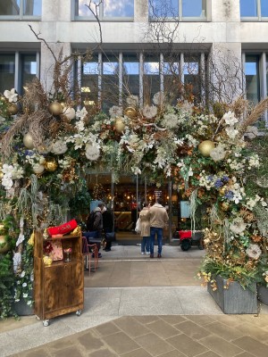 From street, facing venue. Opening ahead with planters on the sides. Path after opening to entrance doors on back, with outdoors seating on back.