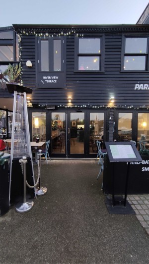 From pavement, facing venue. Opening ahead. Standing sign on right. Restaurant barrier and heater on left.