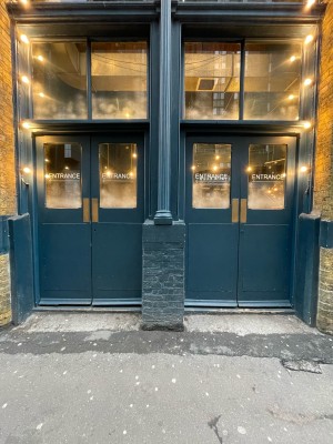 From street facing venue. Two sets of blue wood-panelled glass doors ahead. Pillar in-between doors. No handles. Push at centre to open.