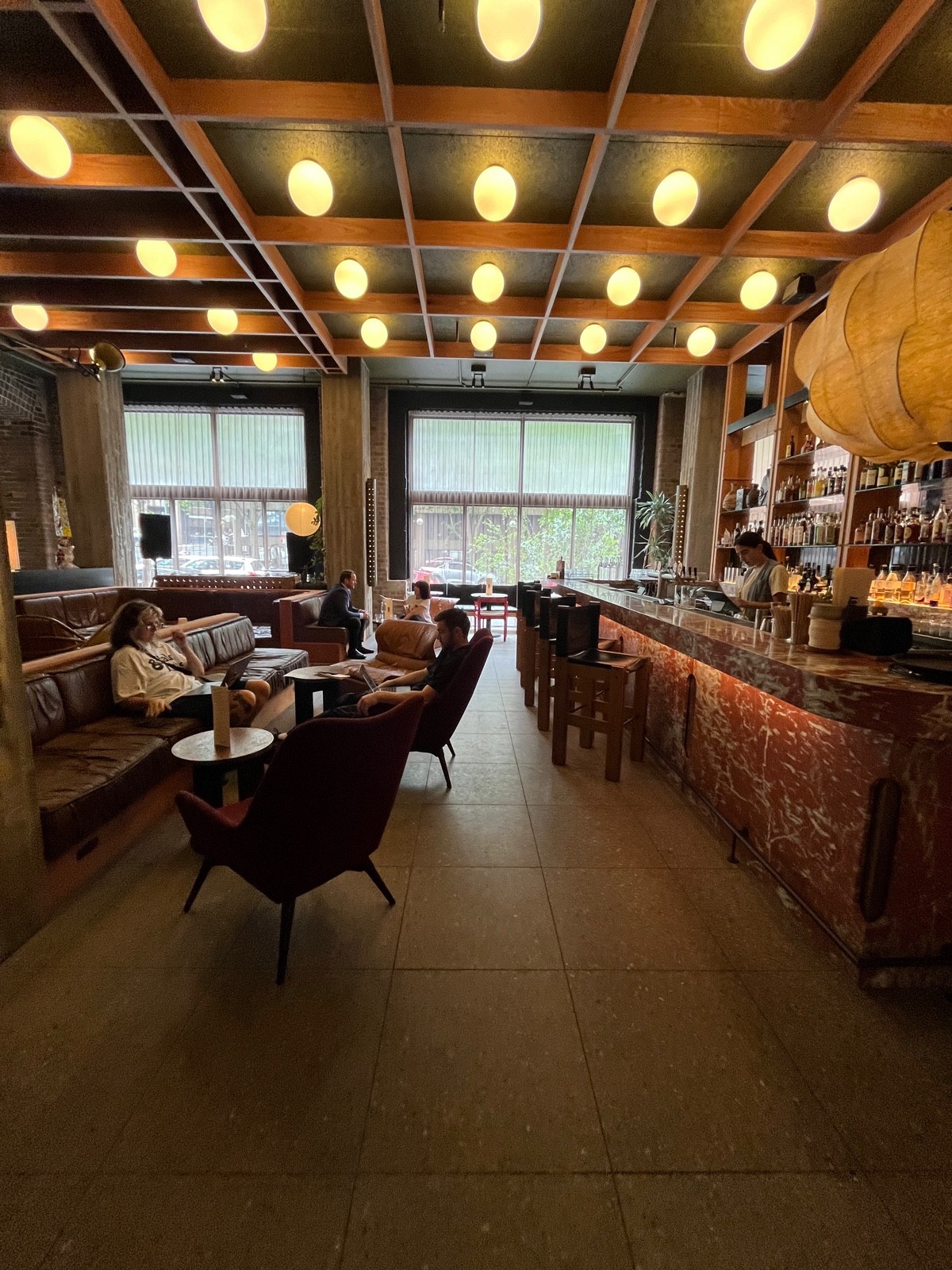 From after armchair facing left. Half booths with low tables and armchairs along left. Bar with backed stools along right. Armchairs obstructing path.