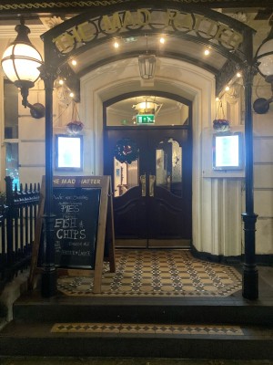 From sidewalk, facing venue. Open archway ahead to entrance on behind. A framed sign on left.