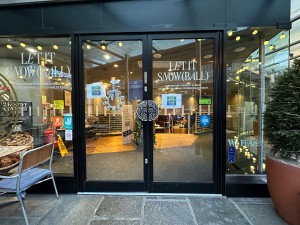Glass doors with black frame. Pull handles in centre to open. Blue doorbell to lower right of doors. Plant to right of doors. Seating to left.