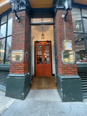 From sidewalk facing venue. Double set of doors. Glass doors with green frame first. Ramp ahead of main doors.