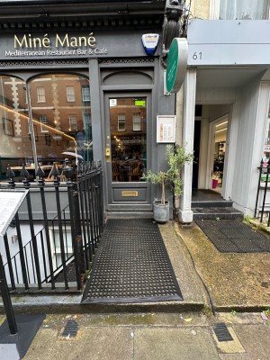 From pavement looking straight at path ahead. Black railing to left. Grey and glass panelled door. Gold handle to left. Push to open.