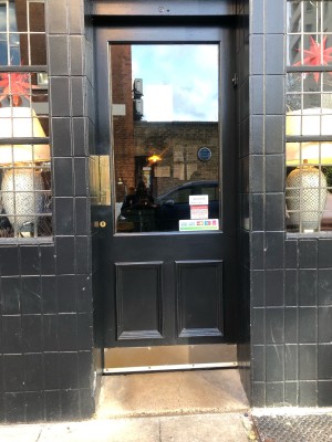 Glass panelled black door. Push on gold panel on left to open.