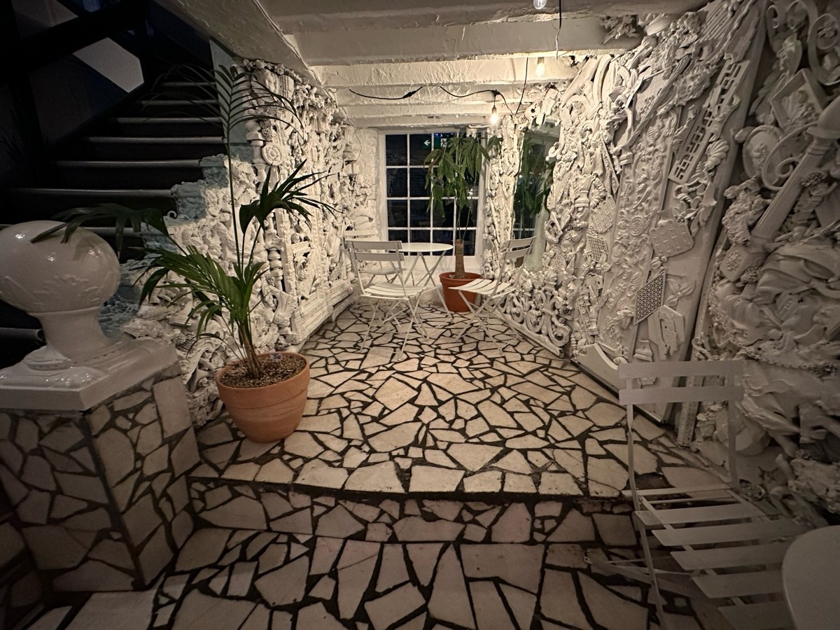 From bottom of stairs facing left. Opening ahead, followed by table and chairs at back. Planter and statue on left.