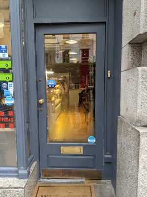 Glass door with grey frame and metal handles. Doorbell to right of door. Push from left to open.