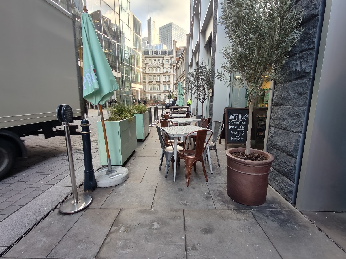 From entrance facing left. Planter on right. Tables and chairs ahead. High table and stools behind. Parasol and restaurant barrier on left.