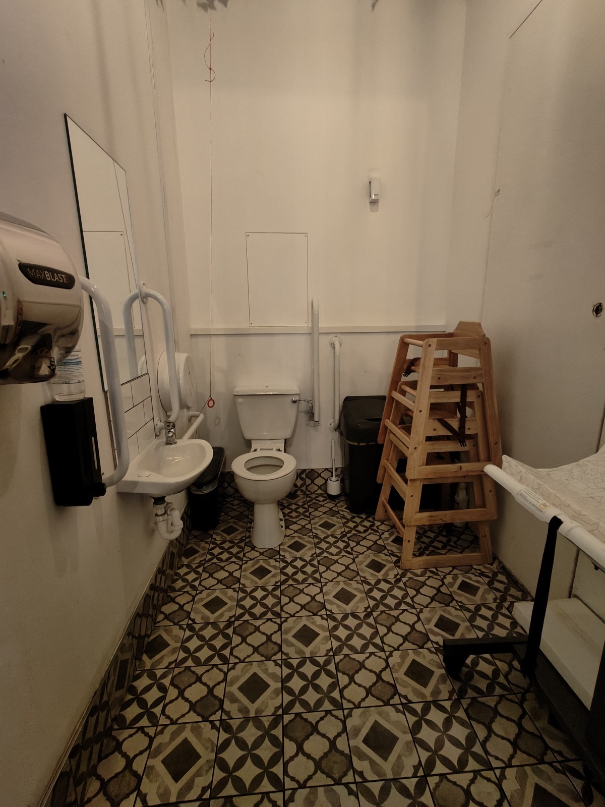 From door facing straight. Sink on left. Toilet at back left. Pull cord to its left. Baby high chairs to its right. Baby change table on right wall.