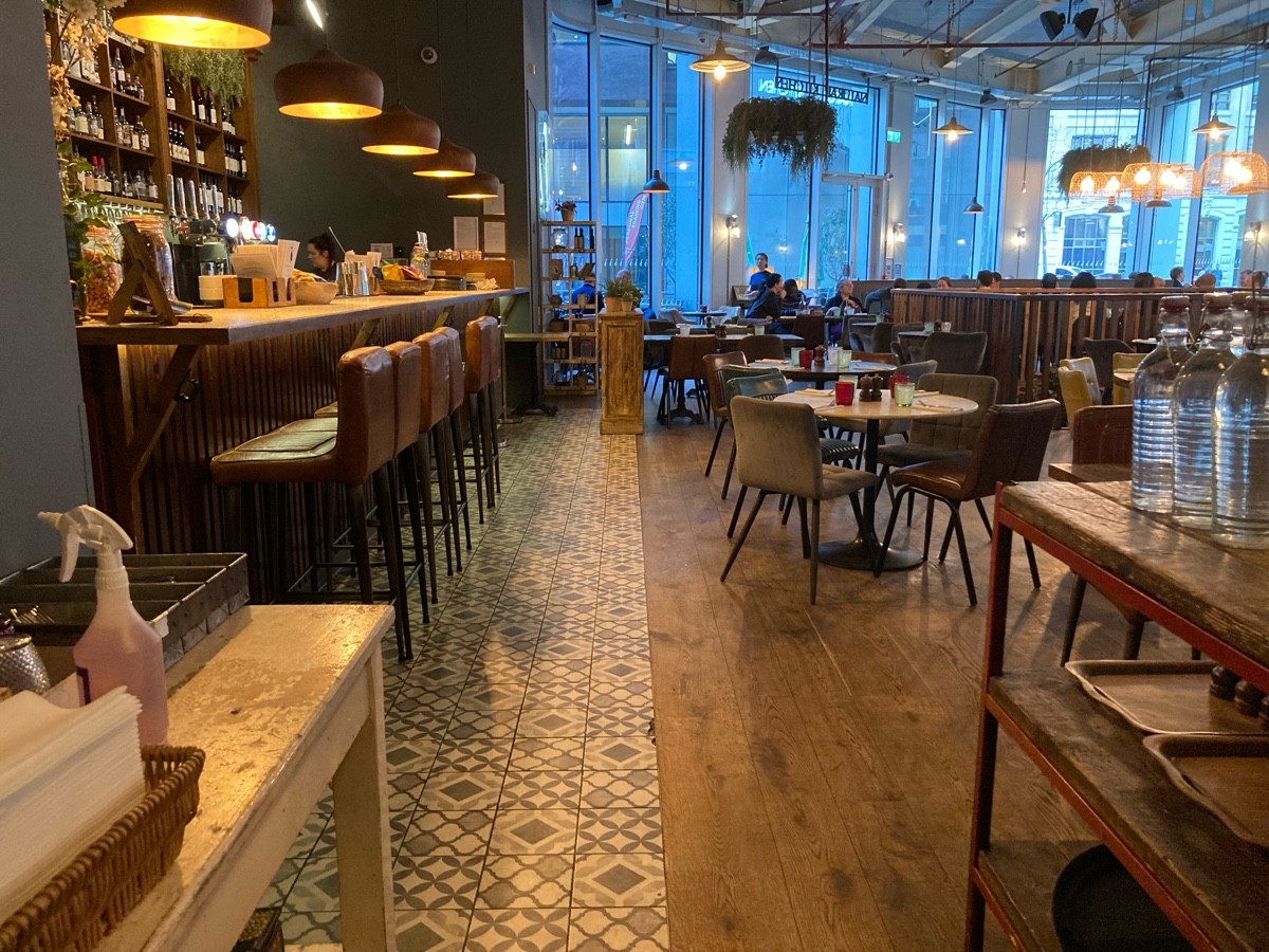 From end of kitchen, facing right. Service desks on left, bar behind. Service desk on right, tables and chairs behind.