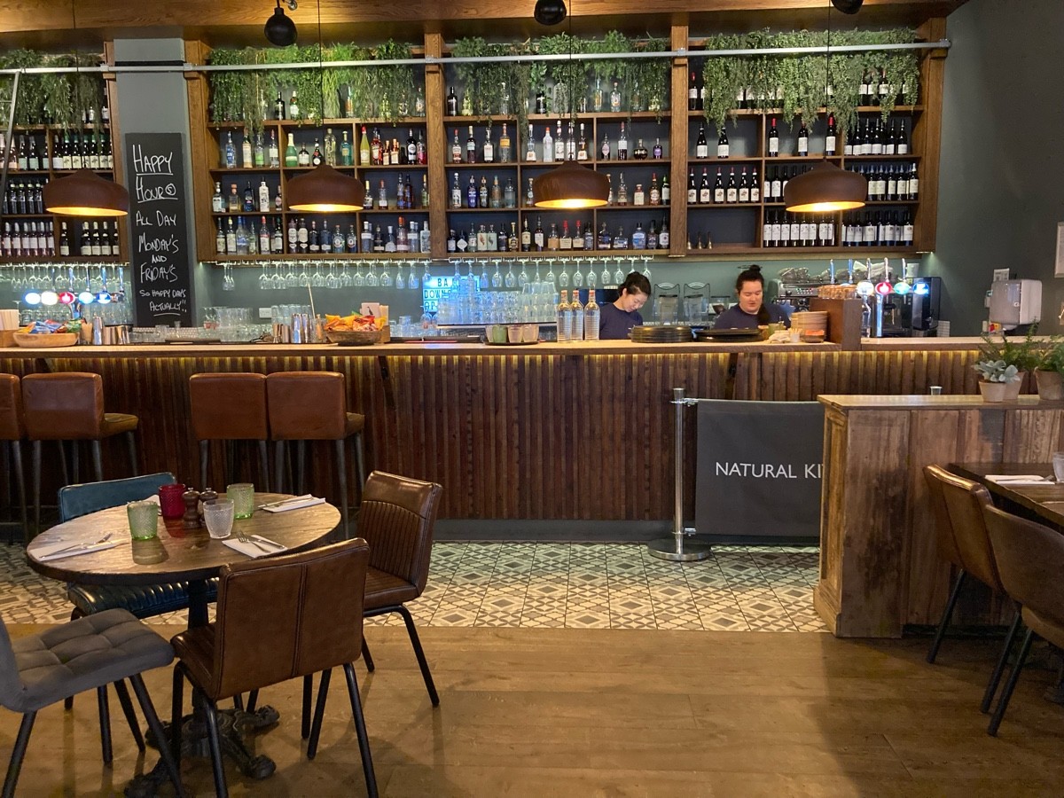 From end of railing, facing straight. Tables and chairs on both sides. Path ahead to bar with stools along back.