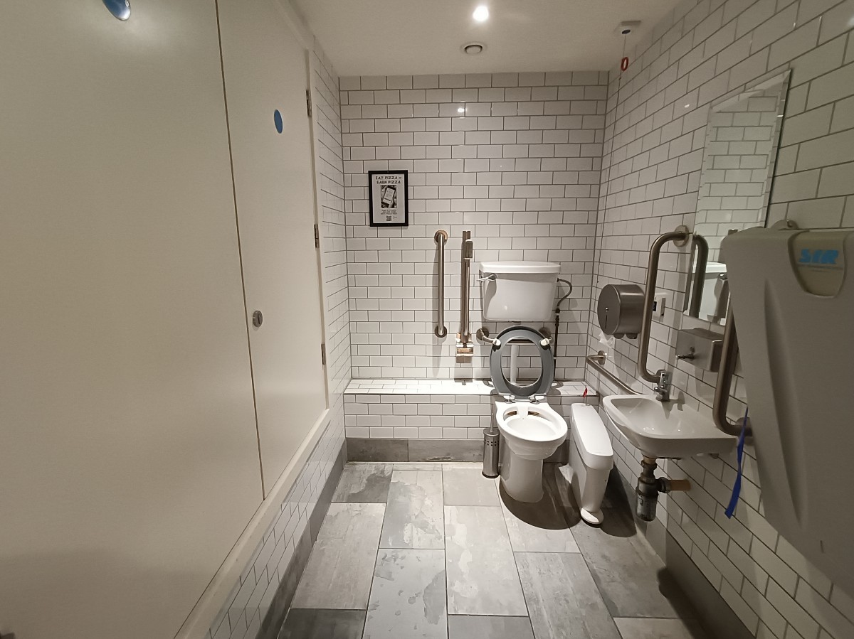 From doorway, looking straight. Baby change on right. Sink behind. Toilet at back right. Emergency pull cord to it's right.