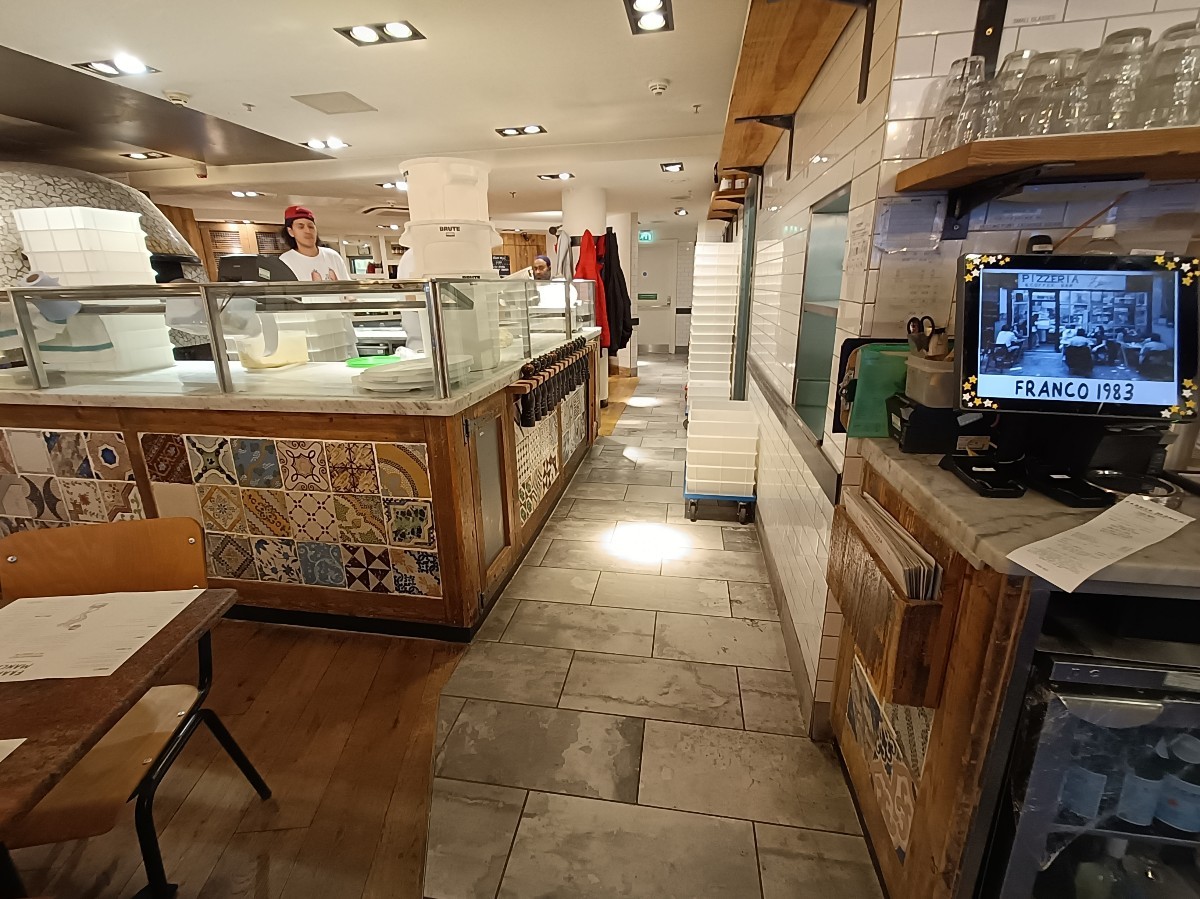 From pillar, looking straight. Tables and chairs on left. Kitchen counter behind. Path in middle to bathroom and side area. Service counters on right.
