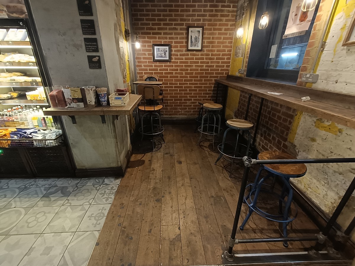 From entrance, looking right. Condiment station and fridges along left. High tables and stools at back and along right.