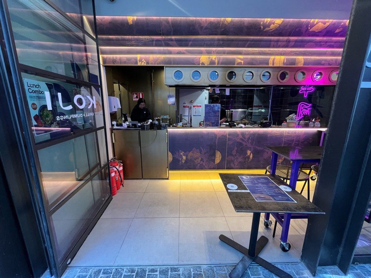 From after outdoor front area facing straight. Glass door with black metal frame slides open to left. Menu table obstructing on right.